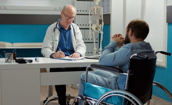 Vecāks ārsts baltā halātā sēž pie galda medicīnas kabinetā un sarunājas ar pacientu ratiņkrēslā. Pacients redzams no mugurpuses, fonā redzams cilvēka skeleta modelis.