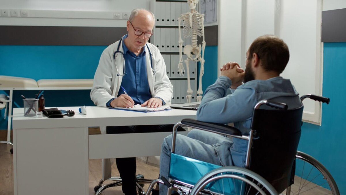 Vecāks ārsts baltā halātā sēž pie galda medicīnas kabinetā un sarunājas ar pacientu ratiņkrēslā. Pacients redzams no mugurpuses, fonā redzams cilvēka skeleta modelis.
